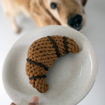 Croissant touwspeeltje op een bord terwijl een hond meekijkt, grappig hondenspeelgoed